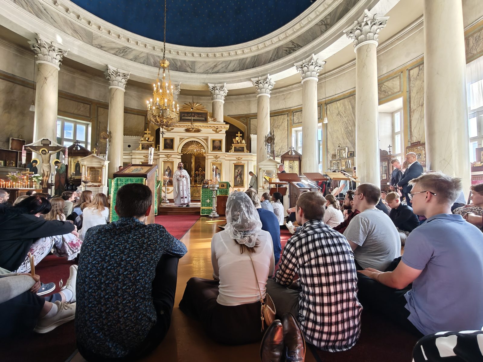 Храм під час проповіді: парафіяни сидять на підлозі спиною до камери й слухають священника, який стоїть перед іконостасом.